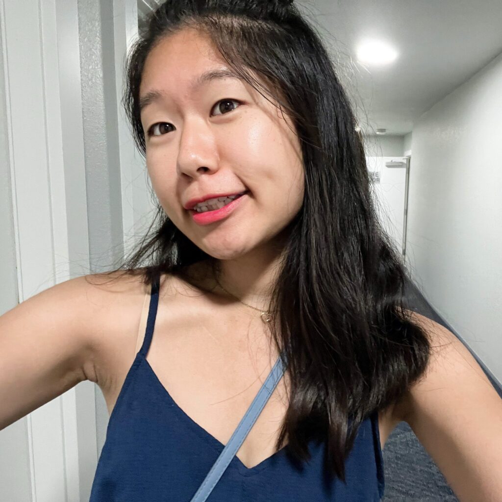A young Asian American woman named Jennifer takes a confident selfie in a well-lit hallway. She wears a navy blue spaghetti strap top, minimal jewelry, and a light blue strap crosses her chest from a shoulder bag. Her long black hair is partially tied up, and she wears red lipstick with a natural makeup look. Her cheerful smile conveys a sense of warmth and authenticity.