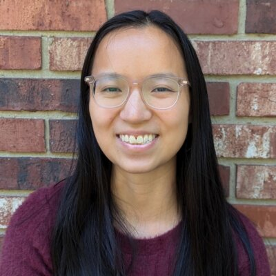 A close-up headshot of Olivia Hall, a young Asian American woman with long black hair and clear glasses. She’s smiling warmly while standing in front of a red and tan brick wall, dressed in a dark maroon sweater.