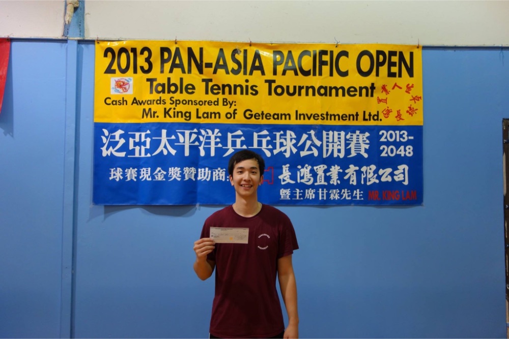 Peter Yau smiles at the camera while holding up a cheque and standing in front of a sign that reads, "2013 Pan-Asia Pacific Open Table Tennis Tournament Cash Awards Sponsored By: Mr. King Lam of Geteam Investment Ltd."