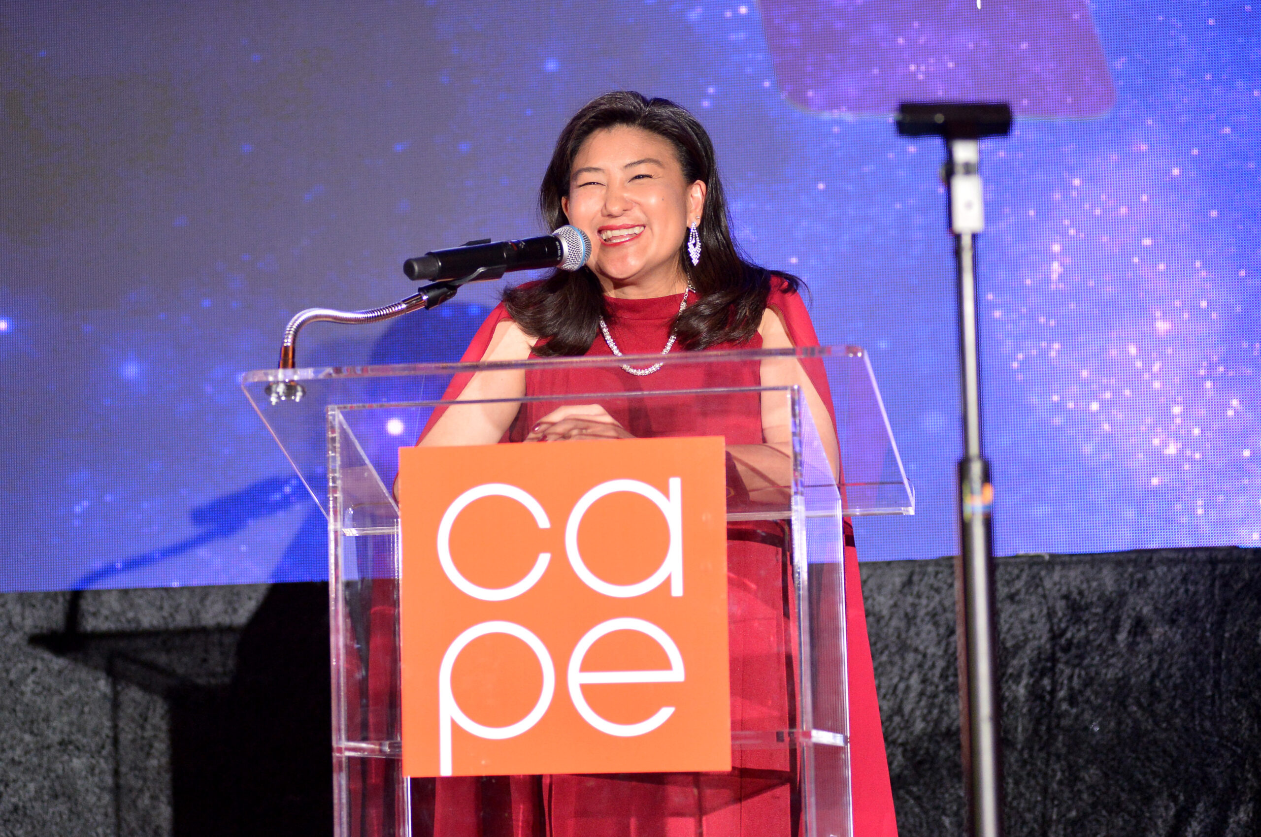 Michelle K. Sugihara standing at podium, smiling, wearing a red dress with cape and silver shiny earrings.