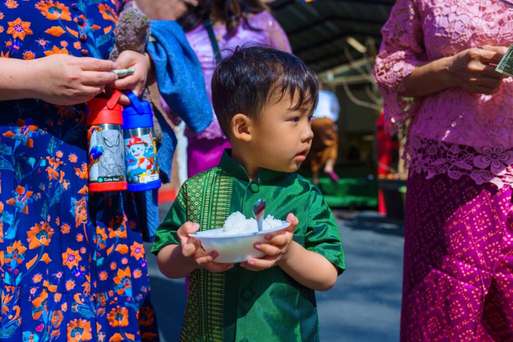 A young boy wearing a shiny green traditional Cambodian outfit holds a bowl of sticky rice while surrounded by people in colorful clothing at a community event.