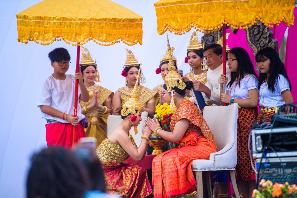 A cultural ceremony with Cambodian traditional dancers in gold outfits and ornate headdresses. One dancer bows respectfully to another seated on stage while being shaded by bright yellow umbrellas.