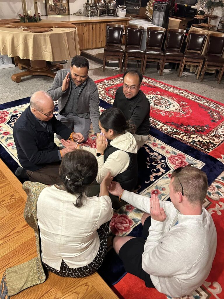 A group photo of Lao American dancers wearing coordinated outfits: women dressed in traditional sinh with bright sashes, and men in white shirts with blue patterned sashes and ties. They pose together outdoors in front of a parade float shaped like an elephant.