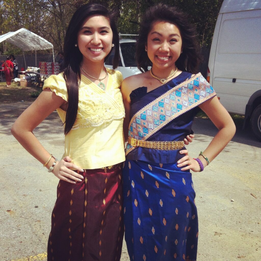 Two young women stand smiling outdoors, dressed in traditional Lao clothing. One wears a yellow blouse with a maroon sinh, while the other wears a dark blue silk sinh with gold embroidery and a blue sash. They stand closely, radiating joy and cultural pride.