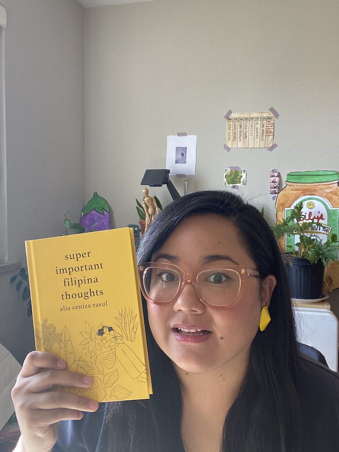 Filipina author Alia Rasul wearing big orange glasses holding a bright yellow book with a slightly perplexed look on her face 
