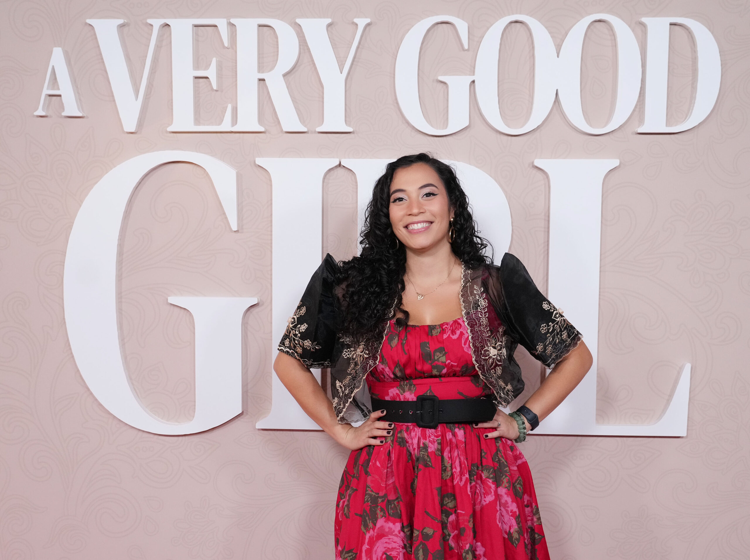 Ashley Rapuano Sanchez stands in front of a blush pink event backdrop that reads “A Very Good Girl.” She wears a vibrant red floral dress paired with a traditional black embroidered bolero. Ashley smiles brightly with her hands on her hips, exuding pride and joy.