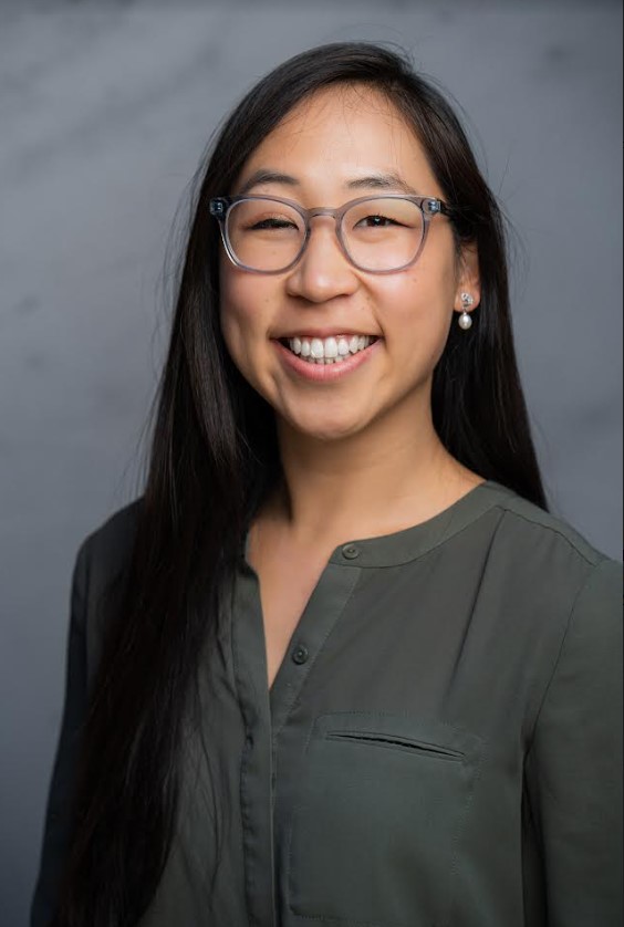 A professional portrait of Angel Wang, a Chinese American woman with long, straight black hair. She is wearing clear-framed glasses, pearl earrings, and a dark green blouse with a pocket detail. Angel is smiling brightly, showcasing her teeth, and looking directly at the camera. The background is a neutral gray with soft lighting, highlighting her face evenly.
