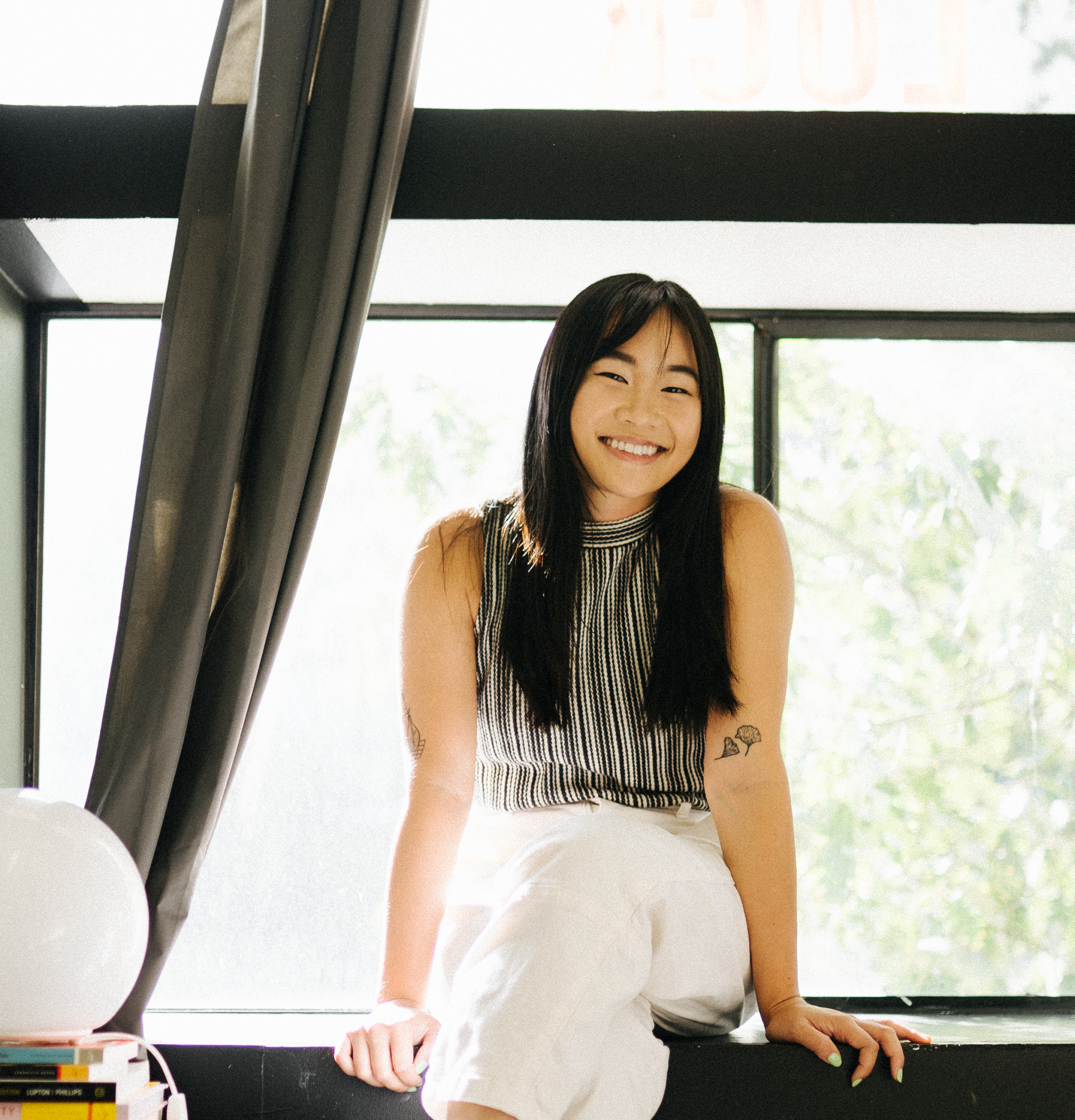 Portrait of Chinese Canadian artist Paige Jung (she/her), smiling and sitting in front of a window. She is wearing a black and white striped tank top with long white pants. Her hair is black and long. She has tattoos near the elbows of each arm. Photo by Solomon Hsu.
