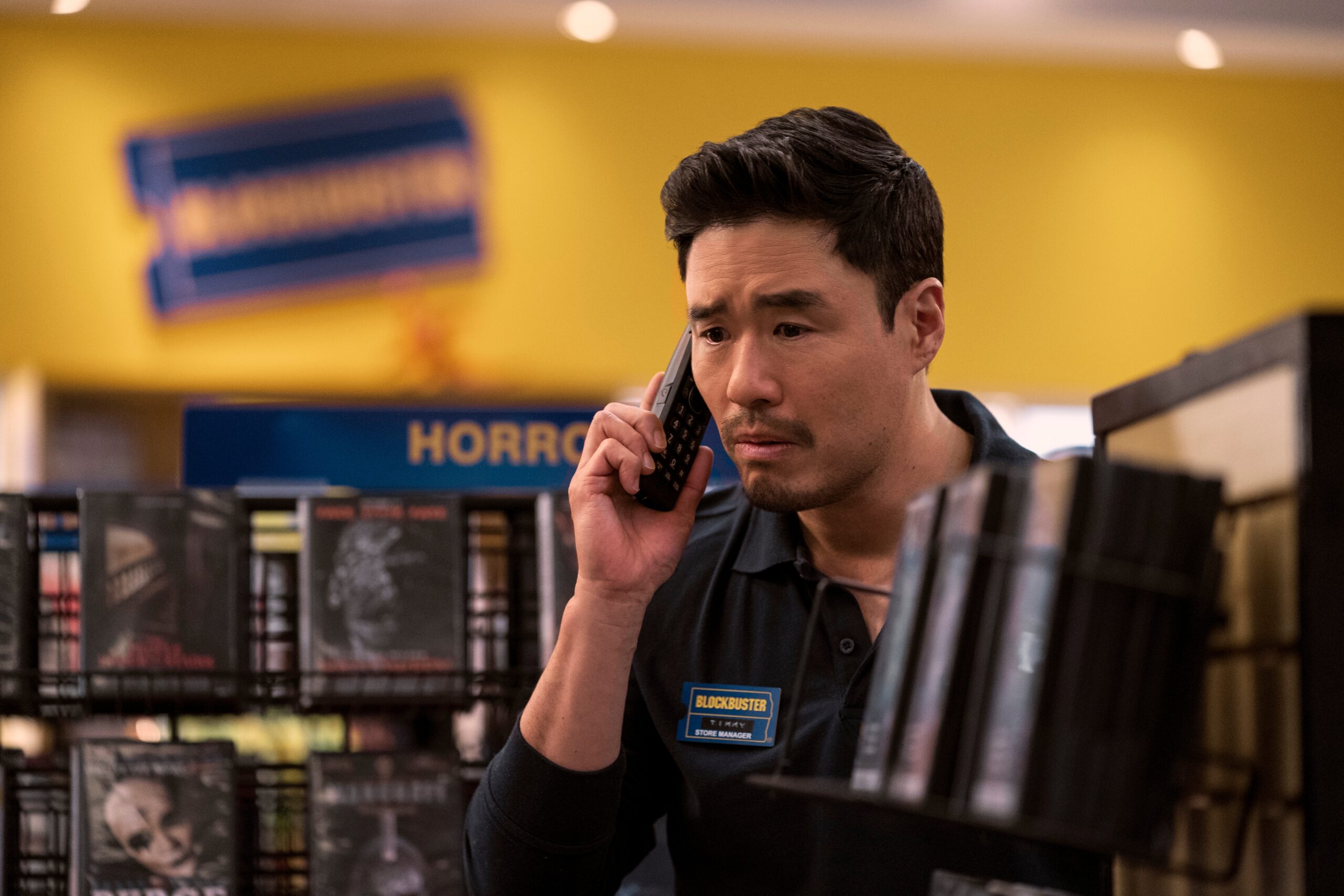 Randall Park as Timmy in episode 101 of Blockbuster. He has a concerned look on his face while holding the phone in between movie shelves. He is standing in front of the horror film section.