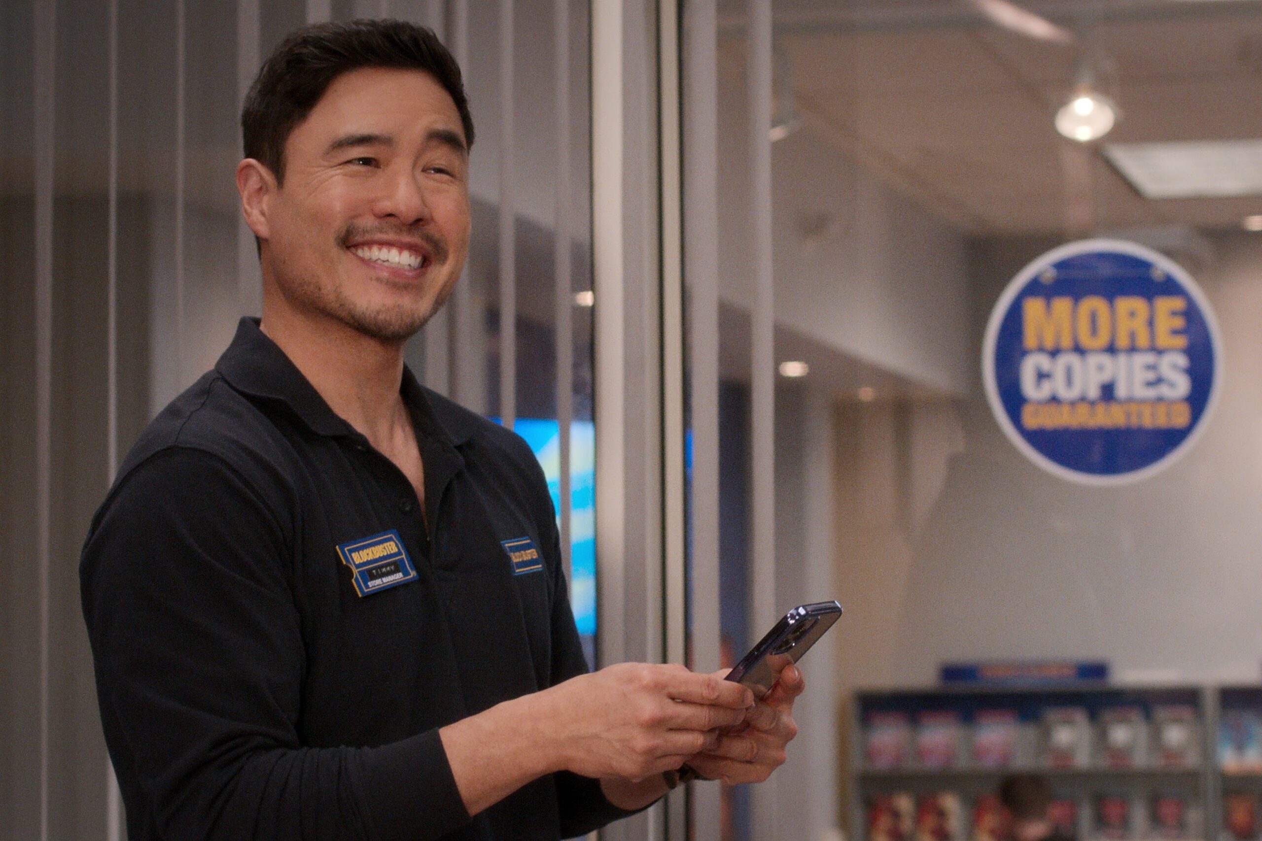 Randall Park smiling while holding a phone on the set of Blockbuster. 