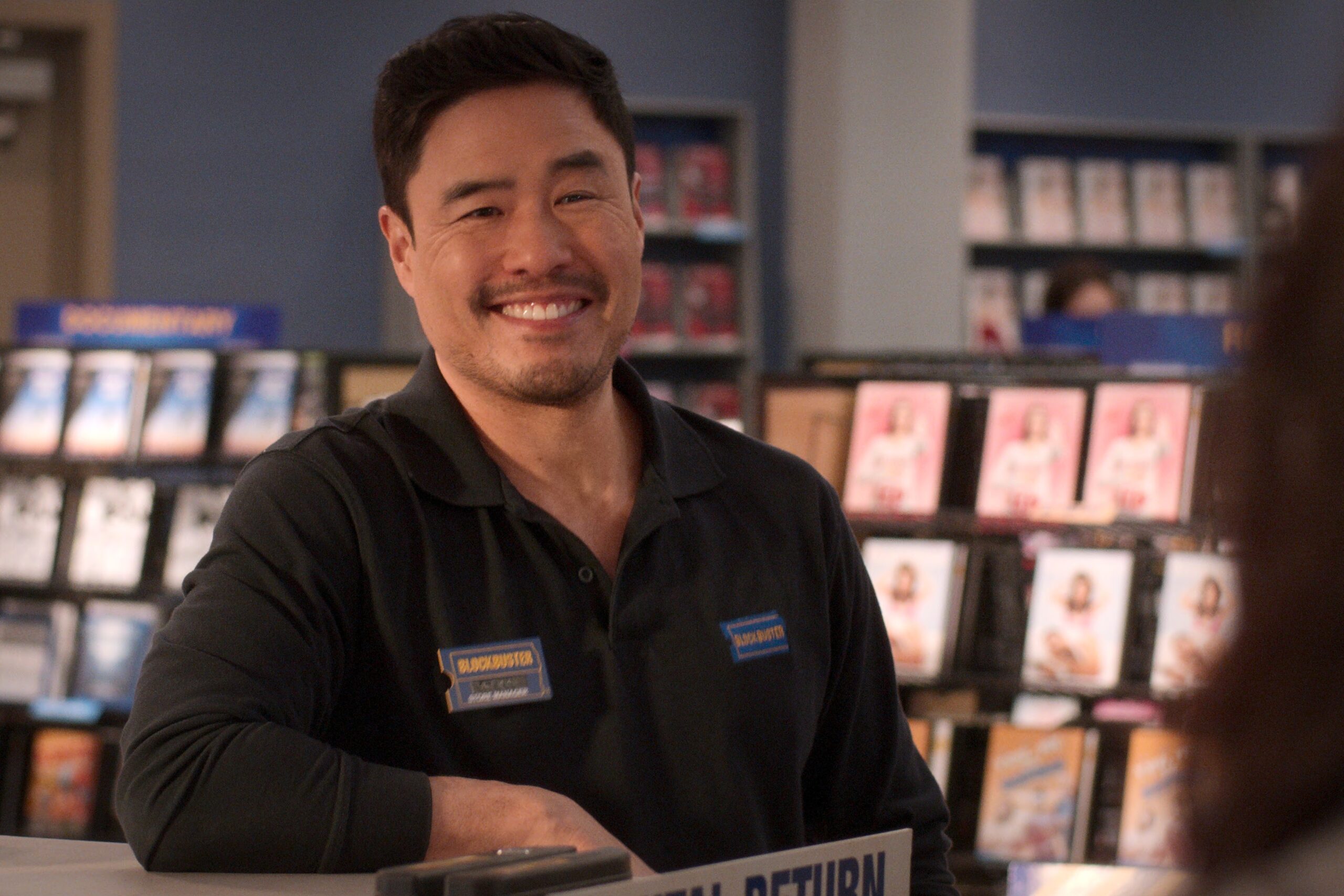 Randall Park smiling as Timmy in episode 106 of Blockbuster. He is leaning against a counter with video shelves behind him. Photo courtesy of Netflix.