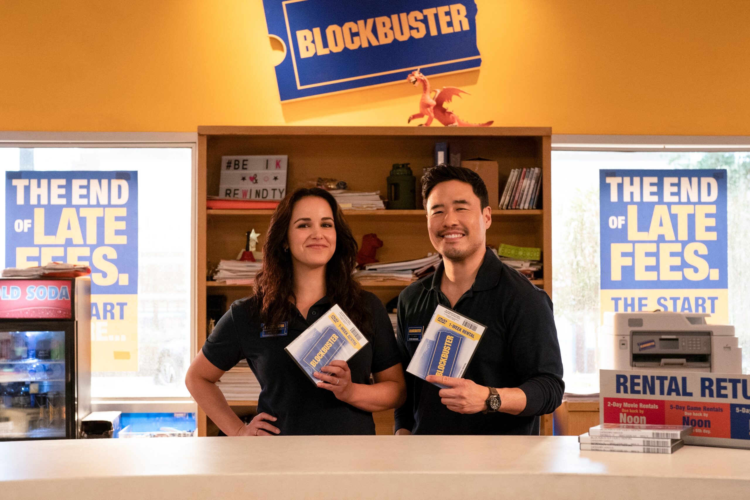 Melissa Fumero as Eliza, Randall Park as Timmy pose while holding Blockbuster DVD's at the front counter of the store.