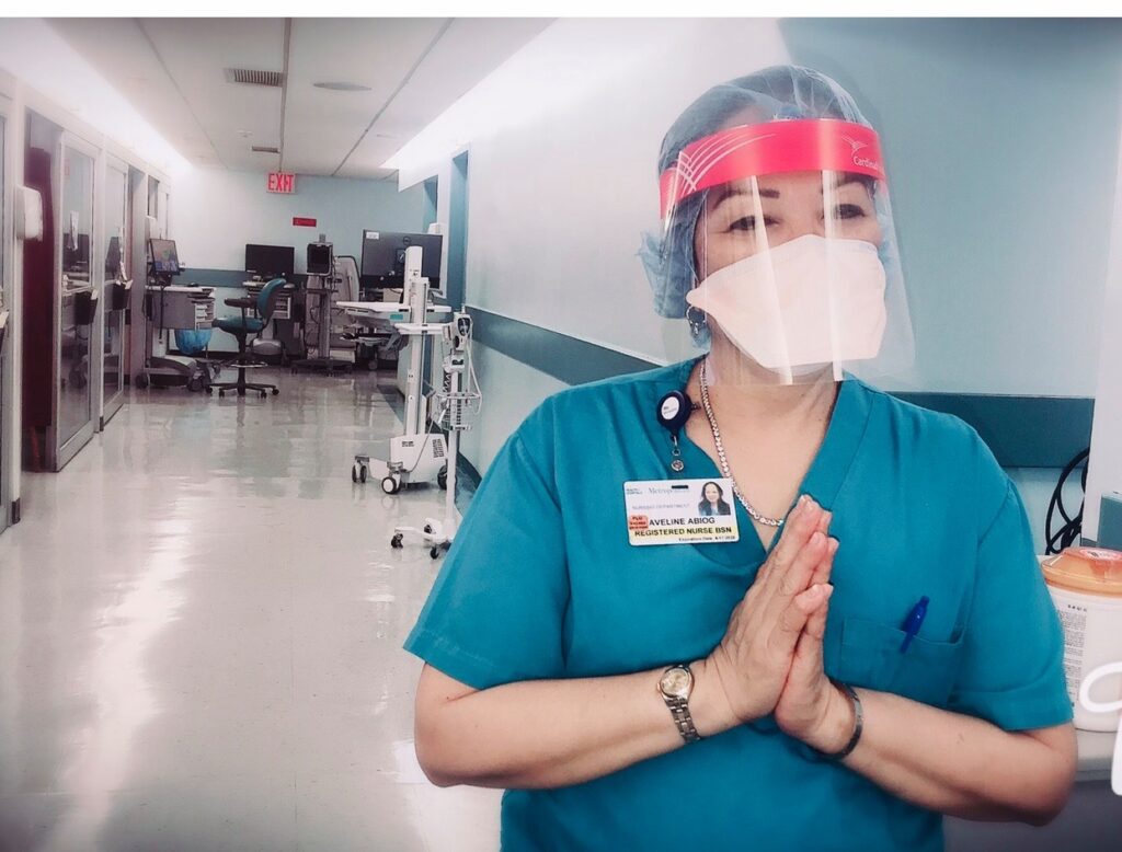 A healthcare worker in blue scrubs, wearing a transparent face shield and a red protective band, standing in a hospital corridor with hands clasped in a gesture of gratitude. The hospital hallway appears quiet and clean.