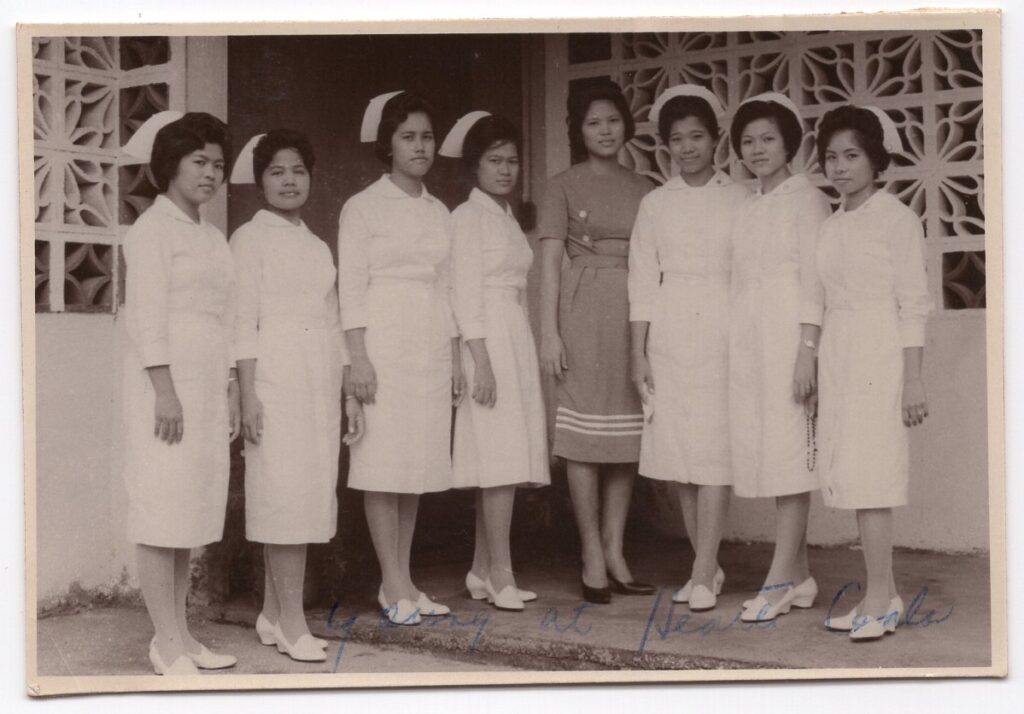 A vintage black-and-white photo of a group of Filipina nurses wearing white uniforms and caps, standing outside a building with a decorative concrete wall. The women are lined up in a respectful pose, representing historical healthcare workers.