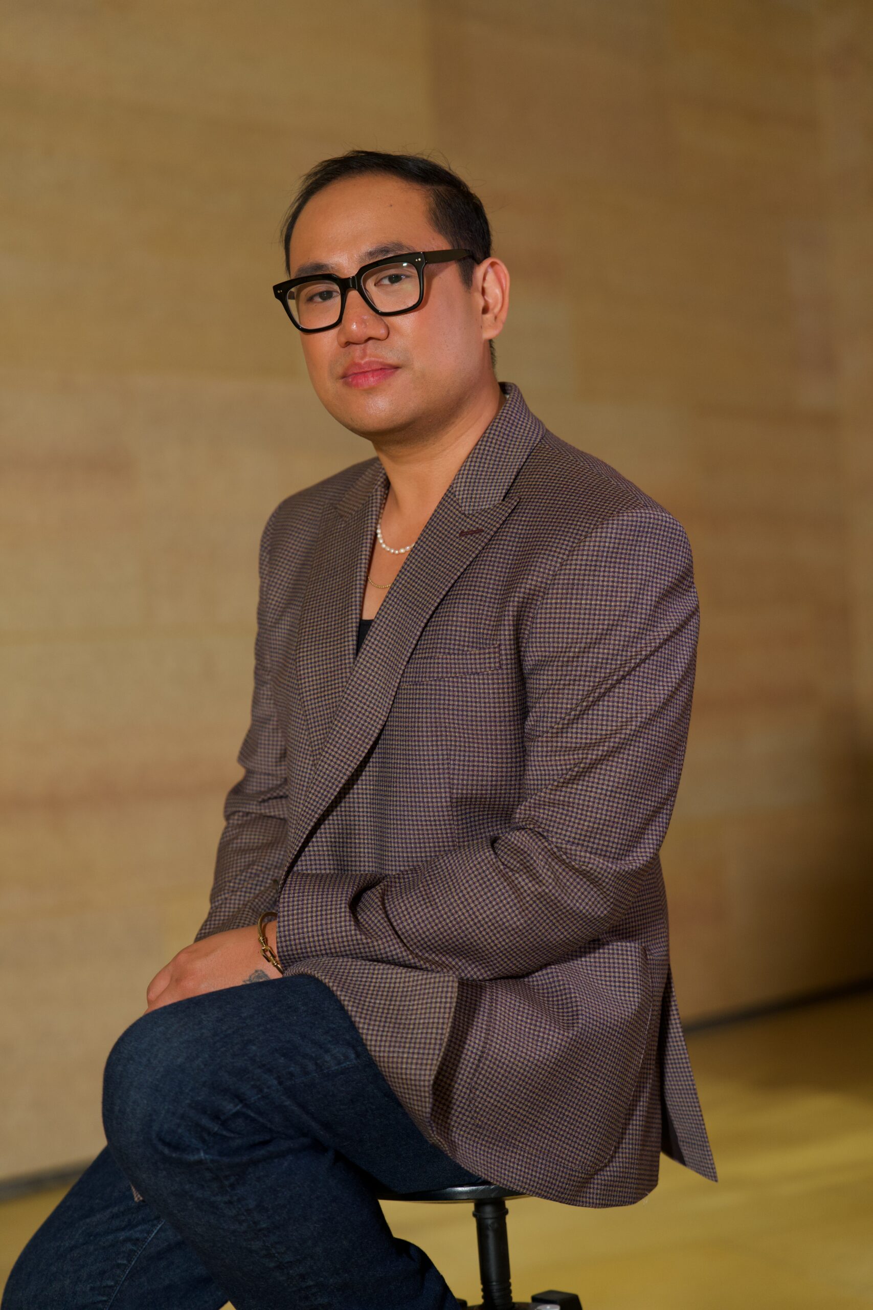 Joseph Carranza, a Filipino American creative, sits confidently on a black stool against a soft beige-toned background. He wears a checkered brown blazer over a black top, paired with dark denim jeans. His look is polished with a pearl necklace, gold bracelet, and black square-framed glasses. He gazes calmly at the camera, exuding a composed and professional presence.
