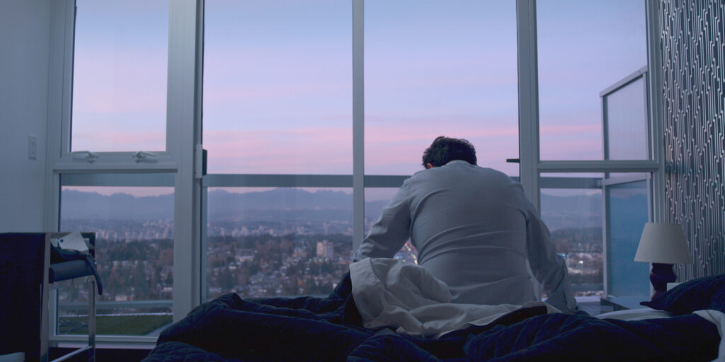 a man in a white shirt sits on a bed with his back to the camera, looking at the floor, facing a window with a view of a city and mountains (Vancouver)