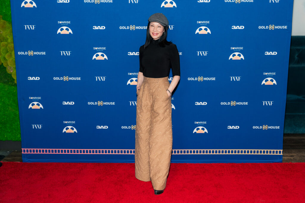 Lucy Liu in a grey beret, black long-sleeved turtleneck and brown pants in front of blue backdrop at Sunrise Collective events