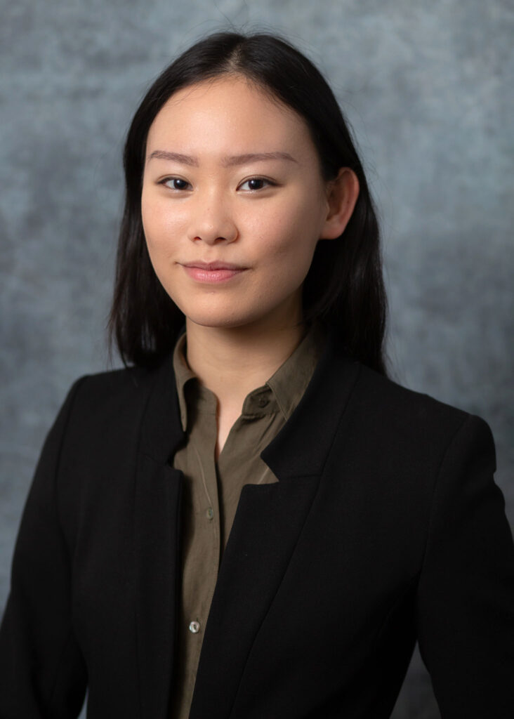 A professional headshot of Elizabeth Sweet, a Korean American adoptee with shoulder-length black hair. She is wearing a black blazer over an olive green button-up shirt. Elizabeth has a poised expression with a slight smile and is looking directly at the camera. The background is a neutral gray with soft, even lighting highlighting her features.