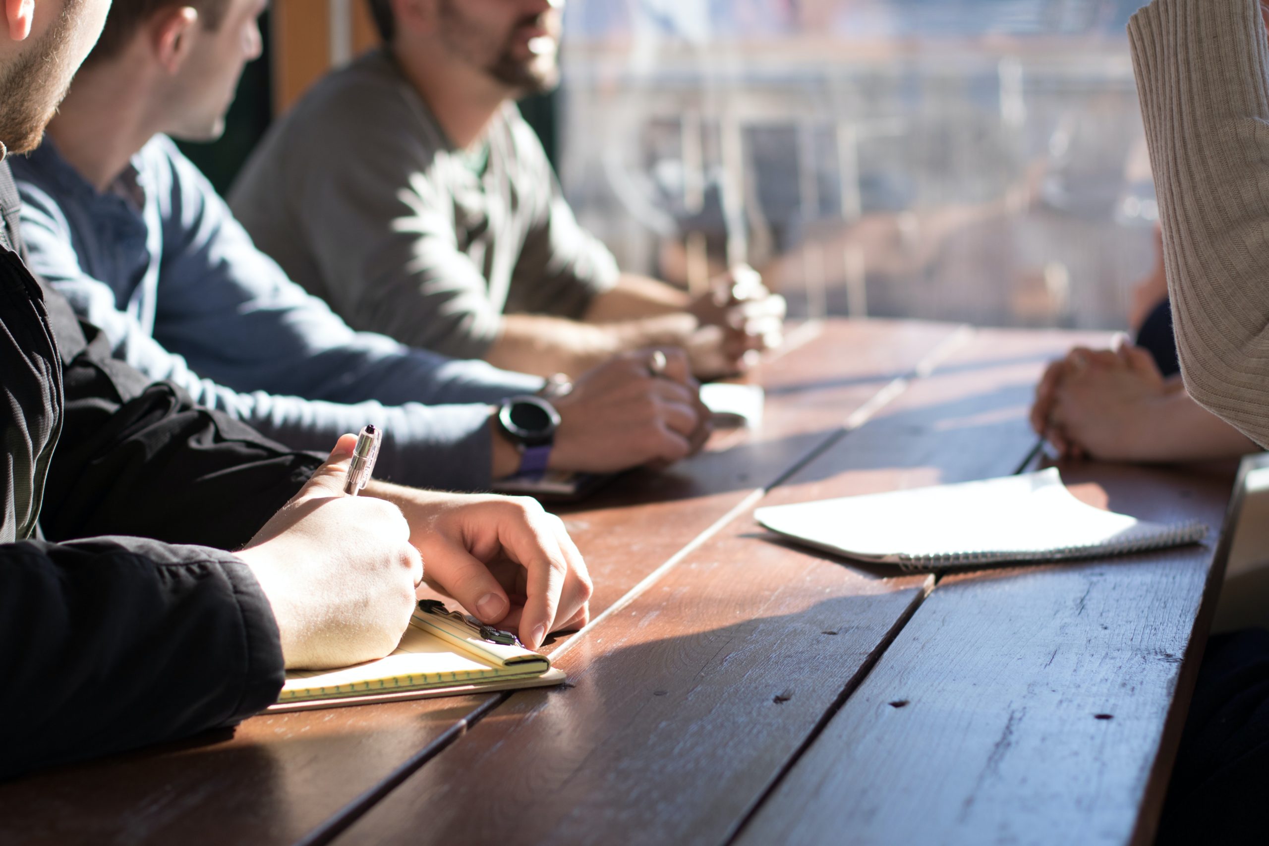 Close up of professionals' seated at a table, with the image focus being their hands and arms. Writing utensils and paper present on table.