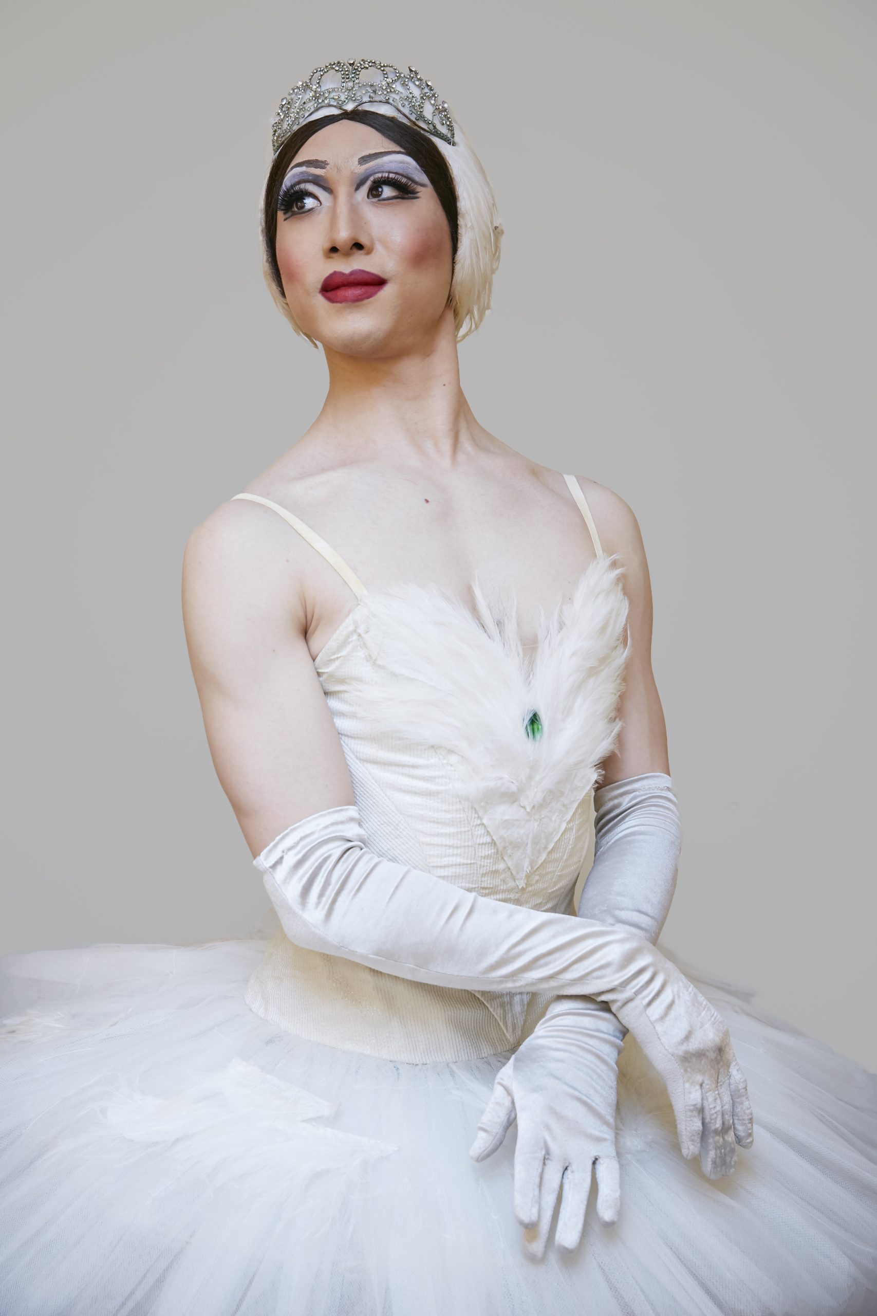 Haojun Xie as Maya Thickenthighya, dressed in a white tutu, with white gloves, complete with show makeup and hair, posed gracefully and smiling into the distance.