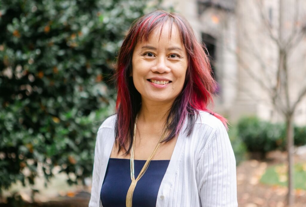 A portrait of Hoang Mai, a Vietnamese American woman, smiling warmly outdoors with a background of green foliage and a blurred cityscape. She has shoulder-length hair with a mix of red and purple highlights. She is wearing a navy blue top, a white open-front cardigan, and a long gold necklace. Her expression is confident and approachable, radiating warmth and positivity.