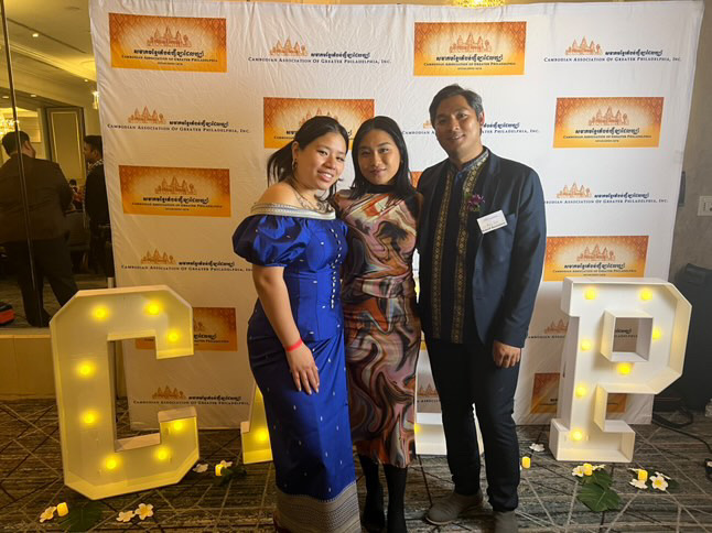 Jenna Lam poses with two friends in front of a Khmer New Year backdrop, wearing a modern swirl-patterned dress, smiling warmly as they celebrate the cultural gathering hosted by the Cambodian Association of Greater Philadelphia.