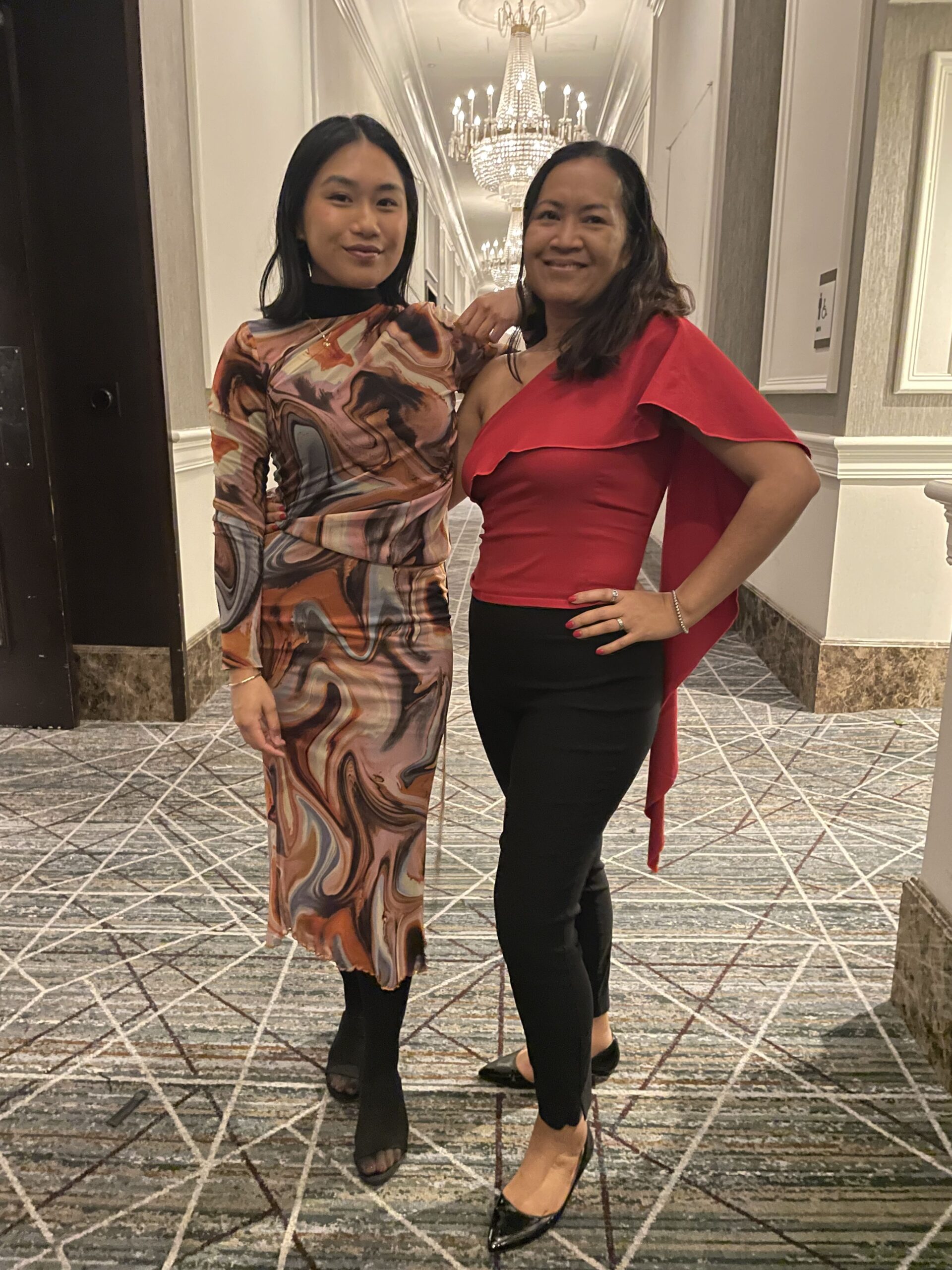 Jenna Lam poses in the hallway of a formal venue with a friend wearing a vibrant red one-shoulder top and black pants, both smiling and dressed up for a Khmer New Year gala or celebration.