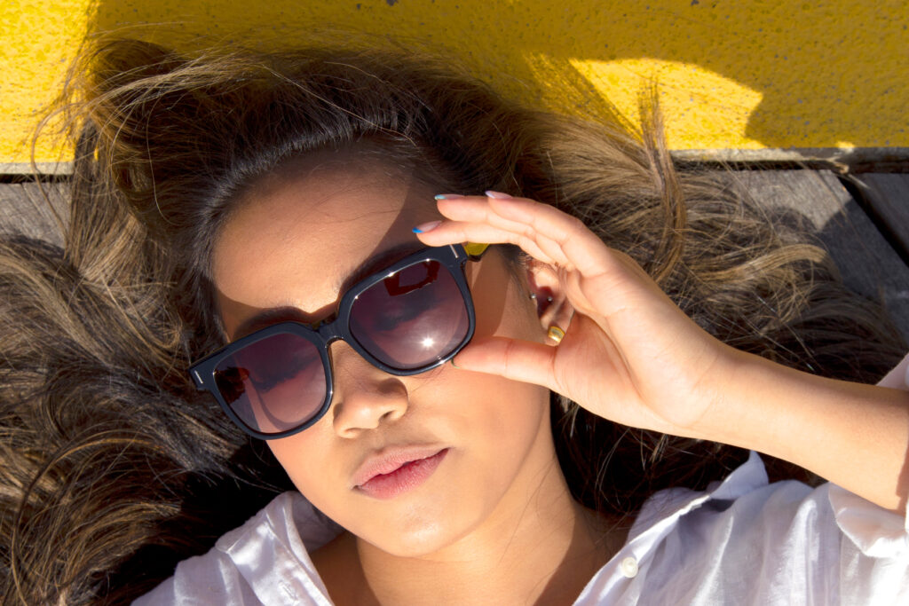 Woman with brown hair lies down and uses black sunglasses to block out the sun. 