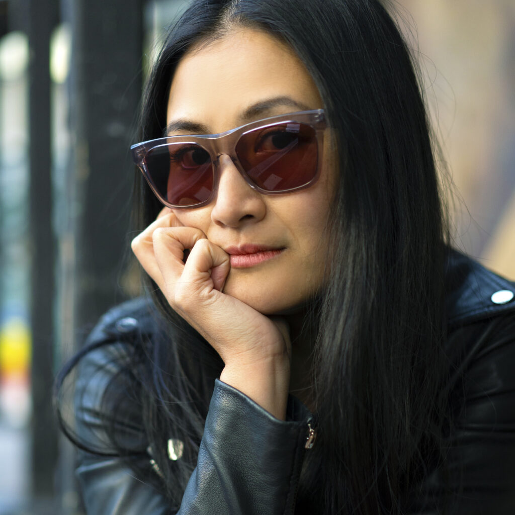 Woman with black hair wears red-tinted sunglasses and stares at the camera. 
