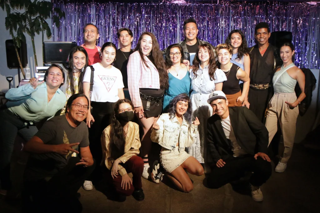Eleven people pose confidently on stage in front of a “Mixed Asian Media” logo. Everyone wears a black or white MAM sweatshirt or hoodie. A few sit on plastic crates, while others stand behind them with crossed arms or casual poses. The lighting casts a cool blue-purple hue across the scene, giving it a polished, editorial look. The group represents a mix of Asian ethnicities and gender expressions, exuding unity and representation.