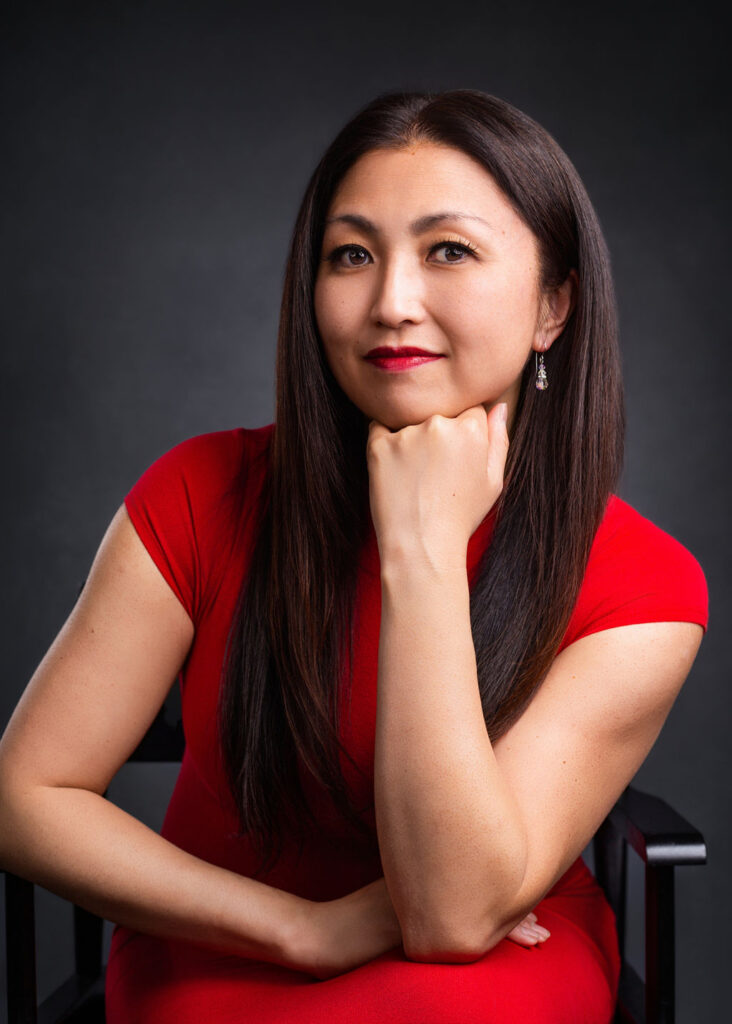 Marisa Hamamoto sits on a black chair wearing a bold red dress. Her long, straight hair falls past her shoulders. She rests her chin on her hand and looks directly at the camera with poise. She wears drop earrings and red lipstick. The portrait is set against a dark grey studio background, conveying a powerful and elegant presence.