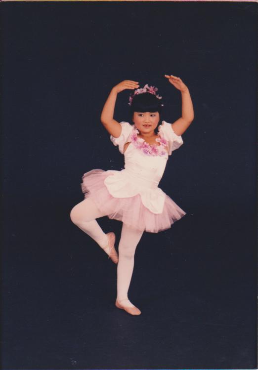A childhood photo of Marisa Hamamoto standing in a ballet pose. She wears a white and pink ballet costume with tulle, floral accents around the neckline, and a flower headpiece. Her arms are raised gracefully above her head, and she balances on one foot with the other leg bent. The background is a dark studio backdrop, highlighting her stance.