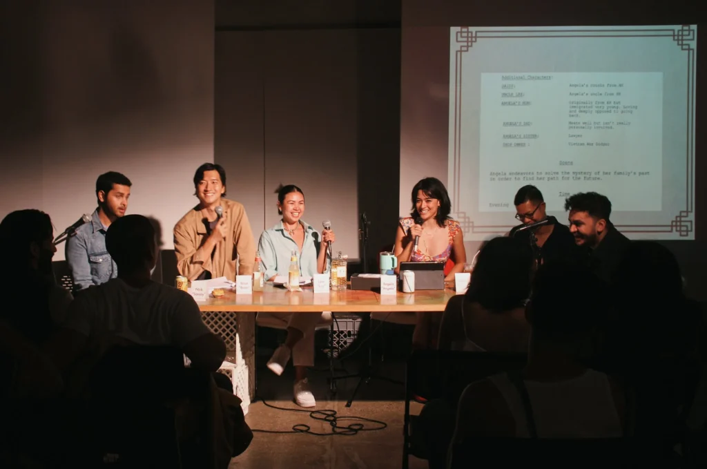 A group of seven Asian creatives sits behind a long table on stage during a live script reading event hosted by Mixed Asian Media. Each person has a microphone and name card in front of them, along with drinks and printed scripts. The room is dimly lit except for warm lighting focused on the panel. The projected screen behind them displays part of a script, titled “Angela’s cousin from NY,” with notes about family dynamics and legacy. The audience is seated in shadow in the foreground, watching attentively. The mood is vibrant and joyful, with panelists laughing, smiling, and engaging the crowd.