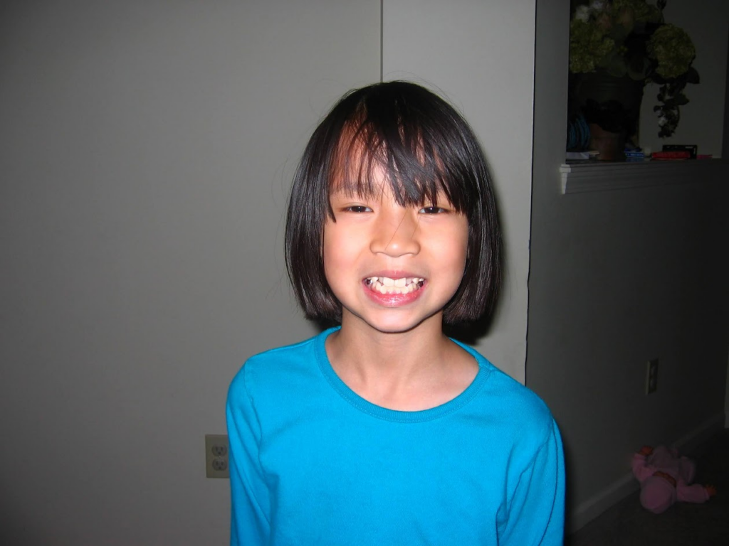 A childhood photo of Olivia Hall smiling with her teeth showing. She wears a bright blue long-sleeve shirt and has a classic bob haircut. The photo is taken indoors against a white wall, with a small plush toy on the floor in the background.