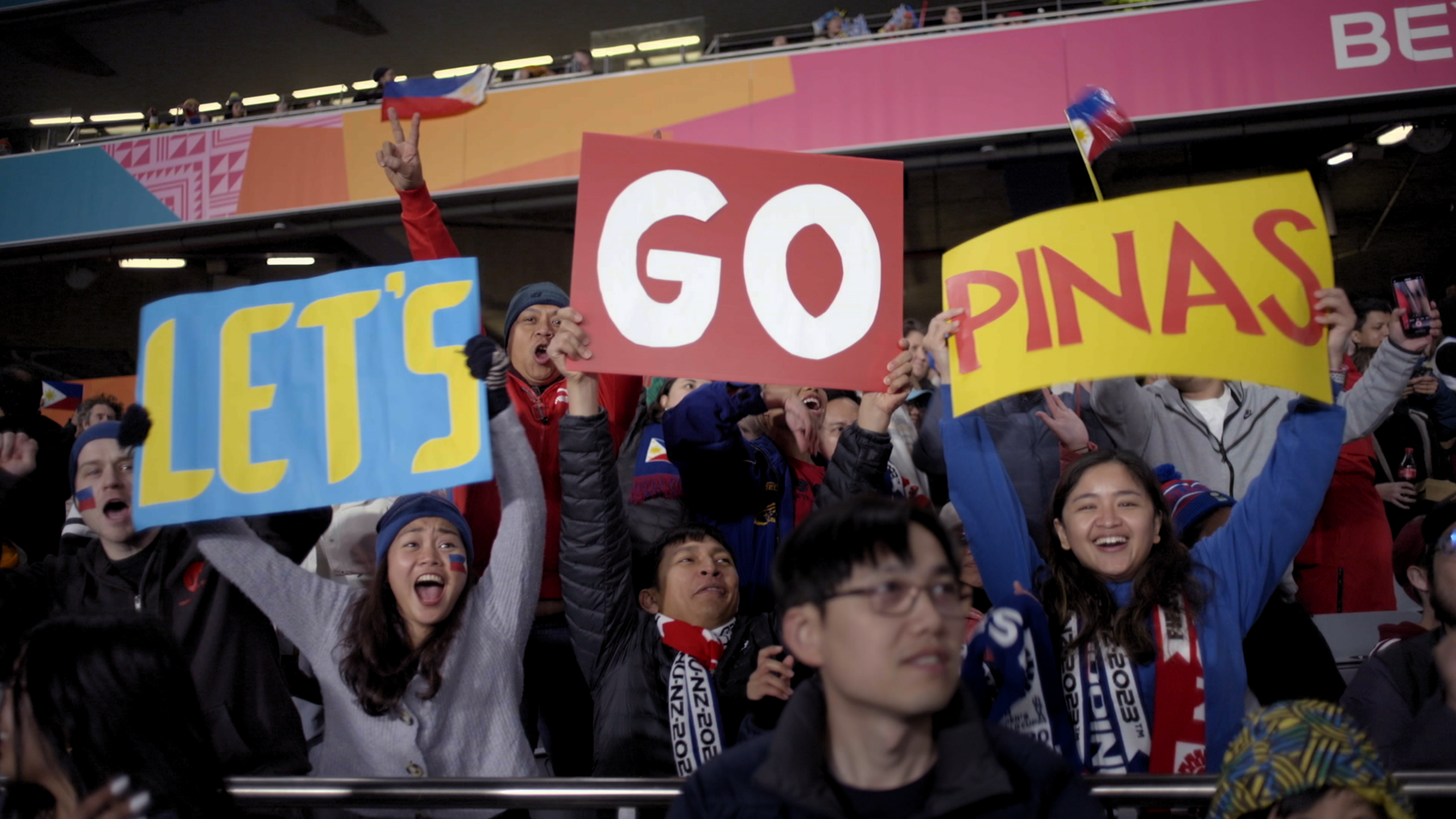 Image of soccer fans in a stadium holding up colorful signs that read "LET'S GO PINAS" while cheering enthusiastically. The atmosphere is vibrant, with fans waving flags and wearing team colors.