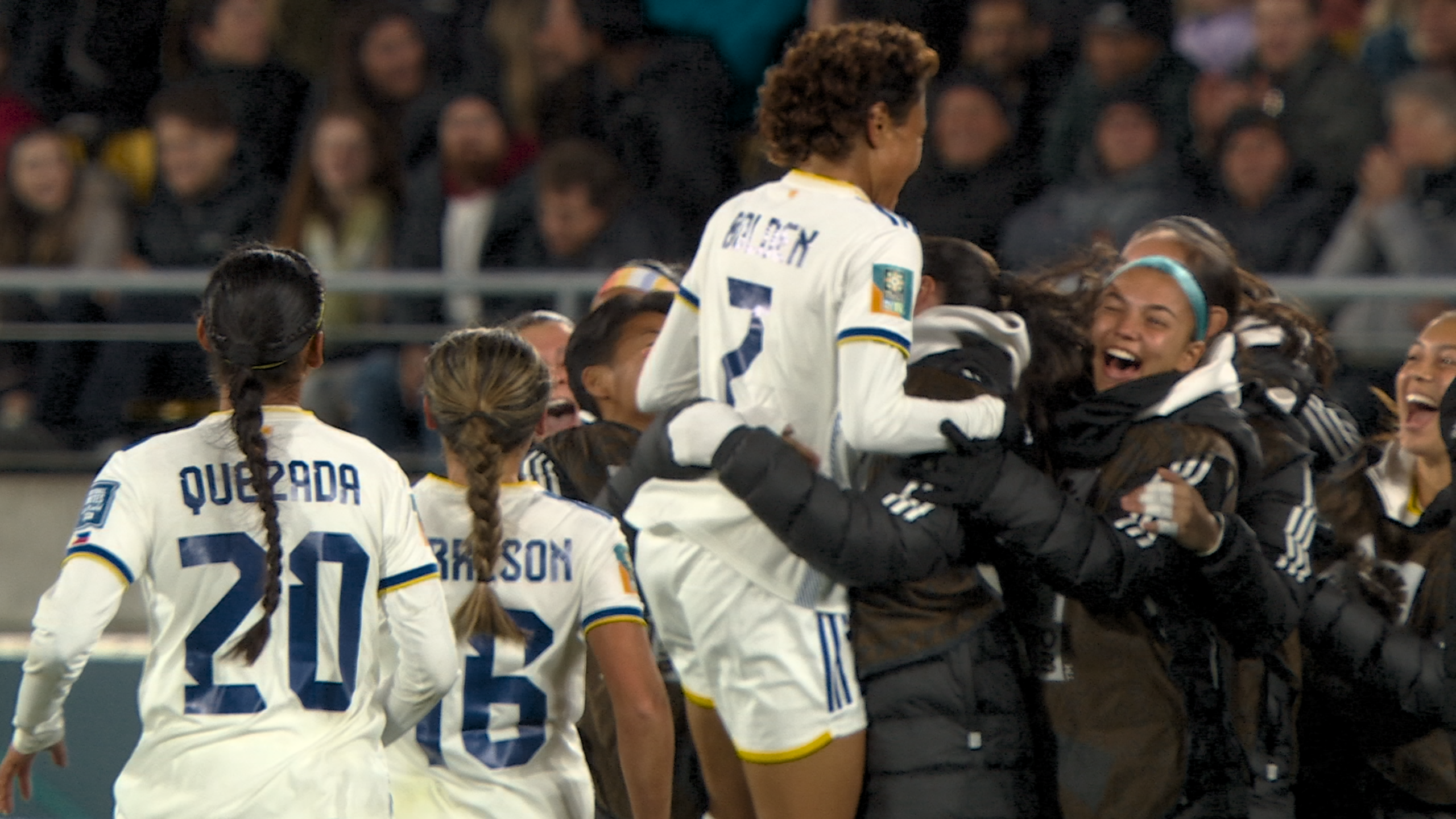 Image of a soccer team celebrating a goal during a match. One player, wearing the number 7 jersey, is lifted in the air while other teammates gather around in excitement. The crowd in the background is cheering.