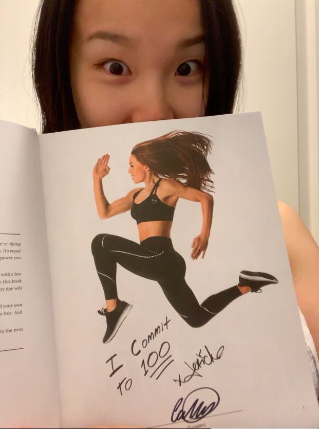 A selfie of the author (Cecilia), the lower half of her face covered by a page that she's holding up. The page features an image of woman in a "stag" jump wearing activewear, and beneath the image are the hand-written words "I commit to 100" and signatures beneath it.