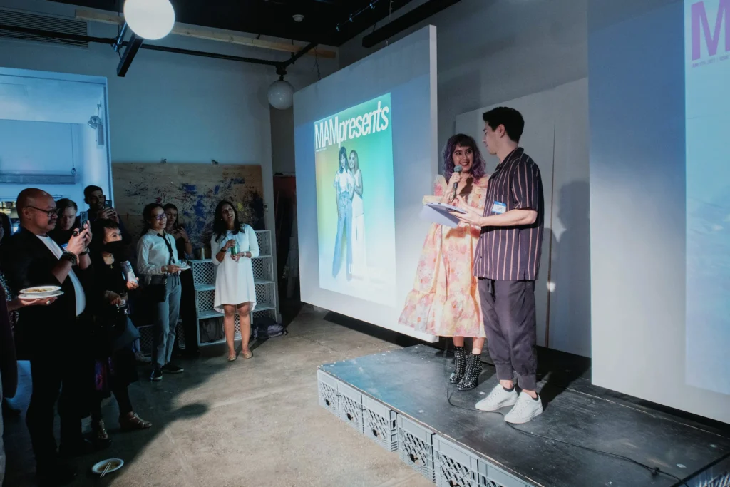 A colorful and lively indoor event shows two presenters standing on a small elevated stage in front of a projected screen displaying the cover of MAM Presents, a Mixed Asian Media publication. The woman on the left wears a long, floral dress and black heeled boots, holding a mic and smiling as she looks at the man beside her, who wears a striped shirt, cropped pants, and sneakers while reading from a notepad. They’re illuminated by soft lighting, while a diverse group of attendees—some holding snacks and drinks—stands closely, watching and recording the presentation with their phones. The atmosphere is casual, joyful, and community-centered.