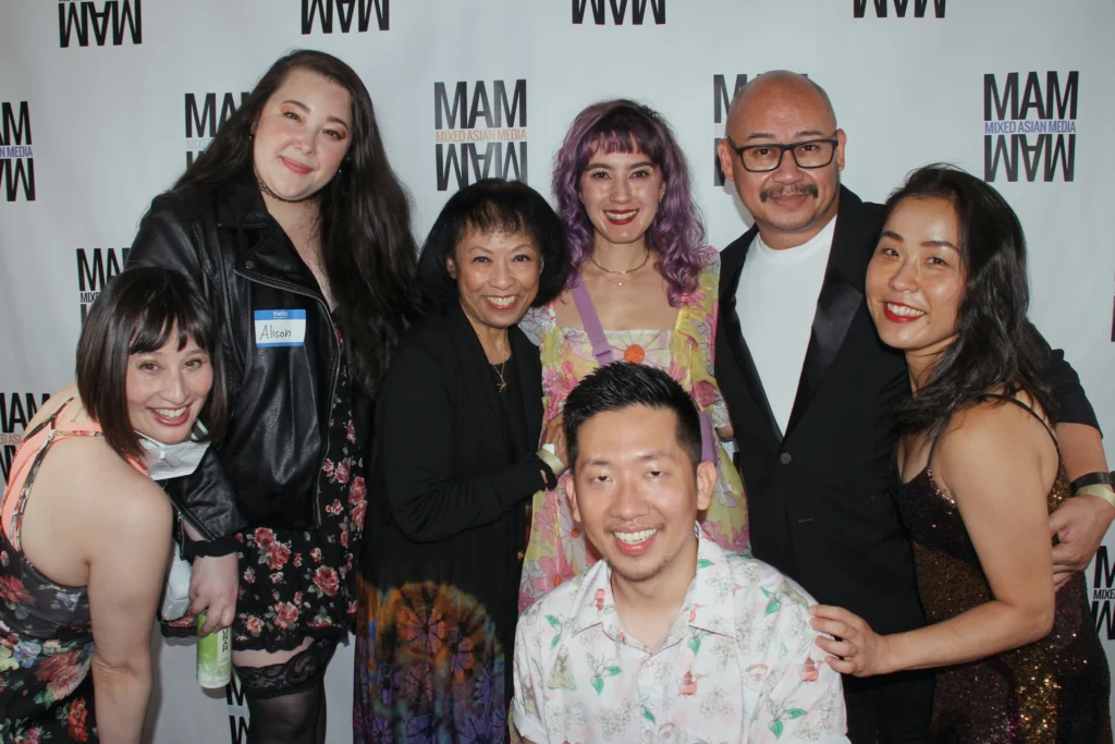 A diverse group of individuals smile warmly at the camera while standing close together in front of a white step-and-repeat backdrop printed with the “MAM” (Mixed Asian Media) logo. Everyone is dressed in vibrant and stylish outfits ranging from floral patterns and leather jackets to formalwear and sequins. The woman in the middle wears a pastel floral dress and has purple hair, standing out brightly. One person kneels in the front center wearing a light floral shirt. The mood is celebratory and full of joy, capturing the inclusive and communal spirit of the event.