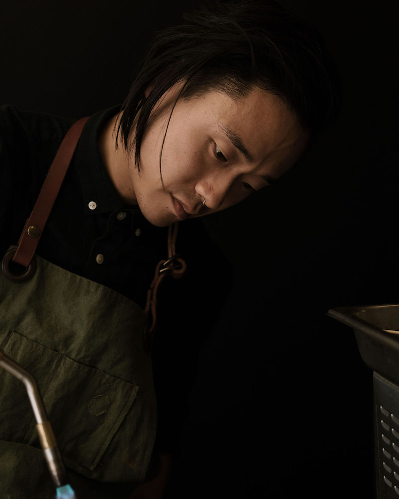 Juno Kim leaning sideways, looking downwards at something. A blowtorch is in the foreground of the image.