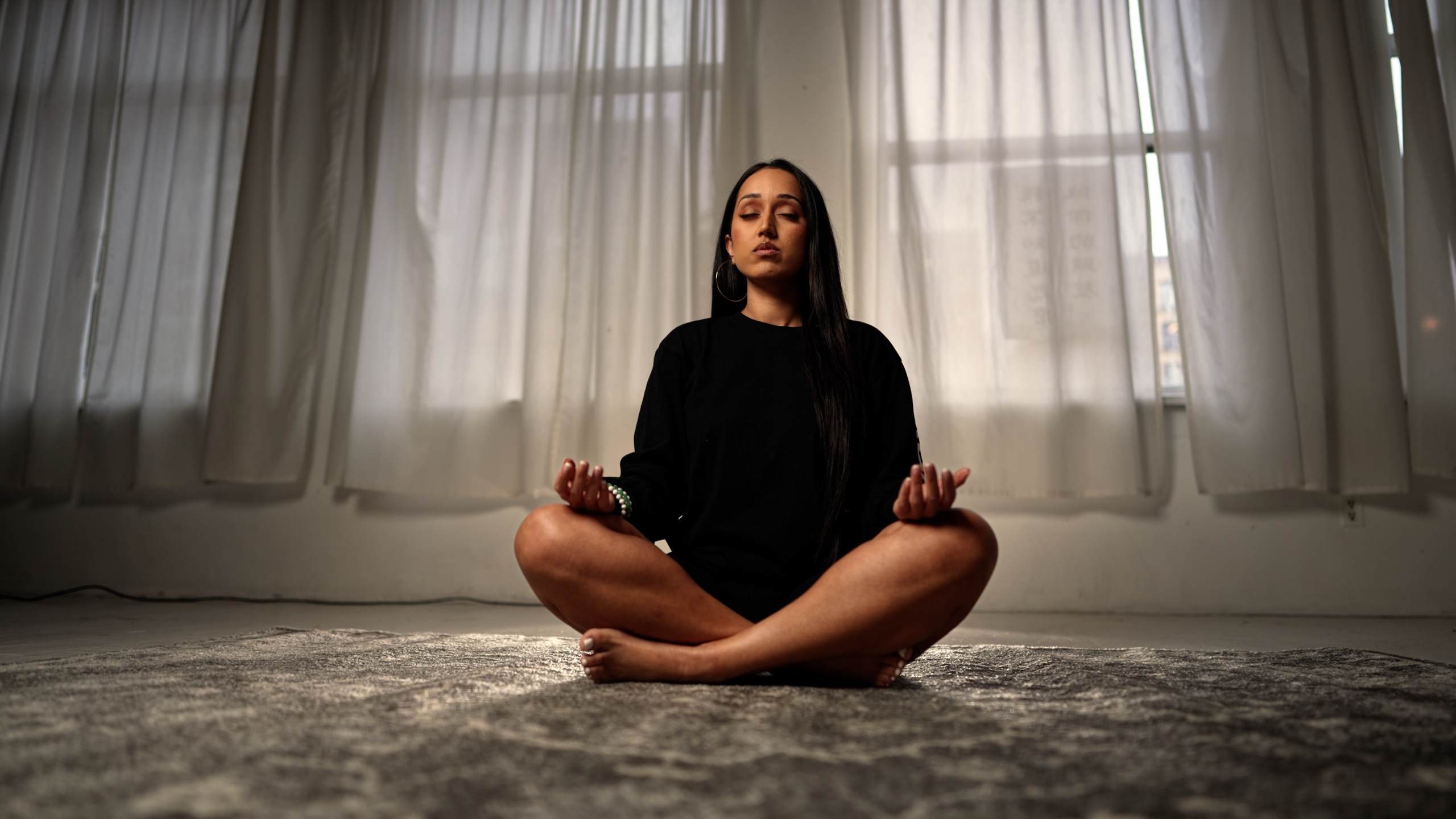 A woman sits crossed legged on the floor with her hands on her knees and with her eyes closed. The wall behind her is curtained and has soft light coming in. 