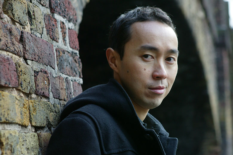 Author Tash Aw standing in front of a brick wall, looking sideways at the camera