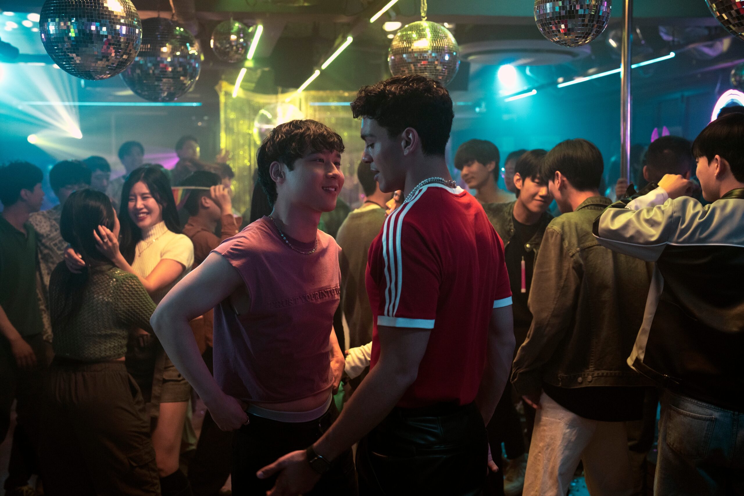 A vibrant nightclub scene with colorful neon lighting and disco balls hanging from the ceiling. In the foreground, two young men stand close together, locking eyes in an intense and possibly flirtatious exchange—one wearing a sleeveless maroon shirt with "Trust Your Intuition" printed on it, and the other in a red Adidas shirt with black leather pants. Around them, partygoers are engaged in conversation and dancing, with some embracing and smiling, creating a lively and dynamic atmosphere.