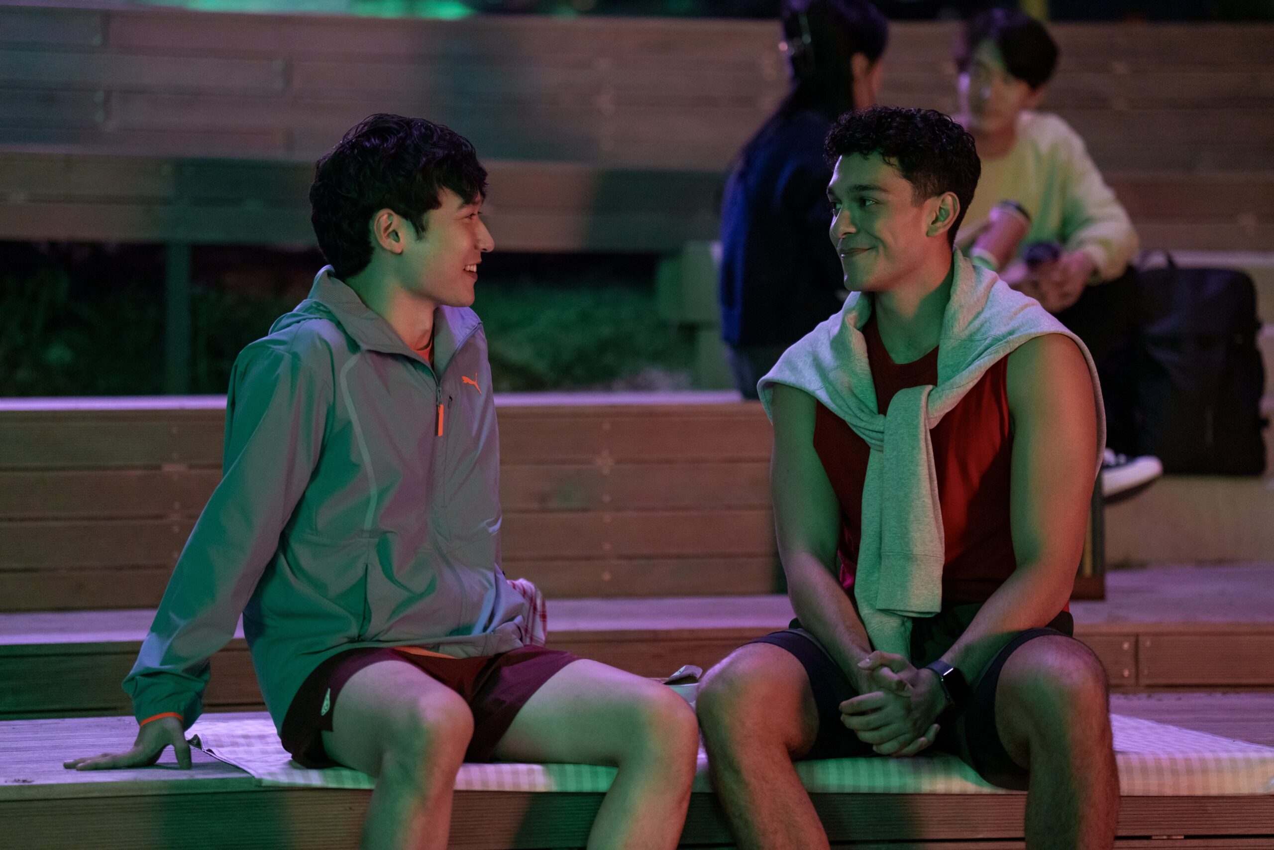 Two young men sit on wooden benches outdoors at night, engaged in a friendly and relaxed conversation. The man on the left is wearing a light gray jacket and maroon shorts, smiling warmly. The man on the right wears a red tank top with a light sweatshirt draped over his shoulders and responds with a soft smile. The scene is illuminated by green and purple lighting, giving it a calm and casual ambiance. In the background, other individuals are sitting and chatting, adding to the social atmosphere.