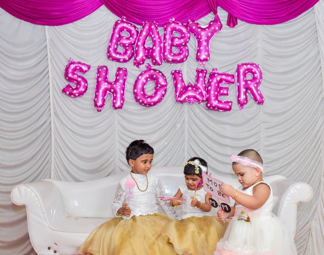 Three Children in yellow dresses sitting on white sofa under pink balloons that spell out "Baby Shower"