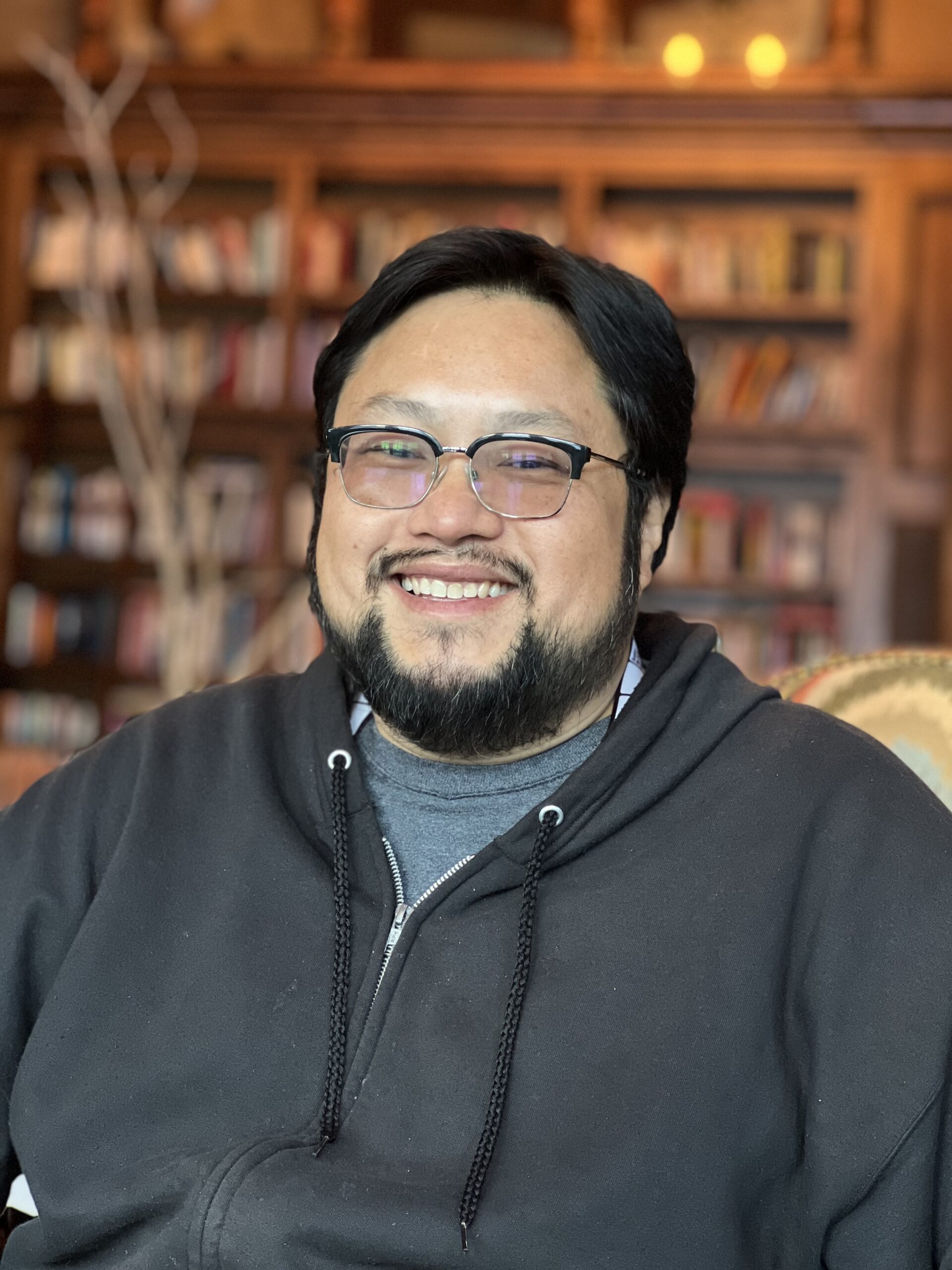 Anderson Le, a Vietnamese American man with short black hair, a beard, and glasses, smiles warmly at the camera. He is wearing a black zip-up hoodie over a grey shirt and is seated in front of a warmly lit, wooden bookcase filled with books.