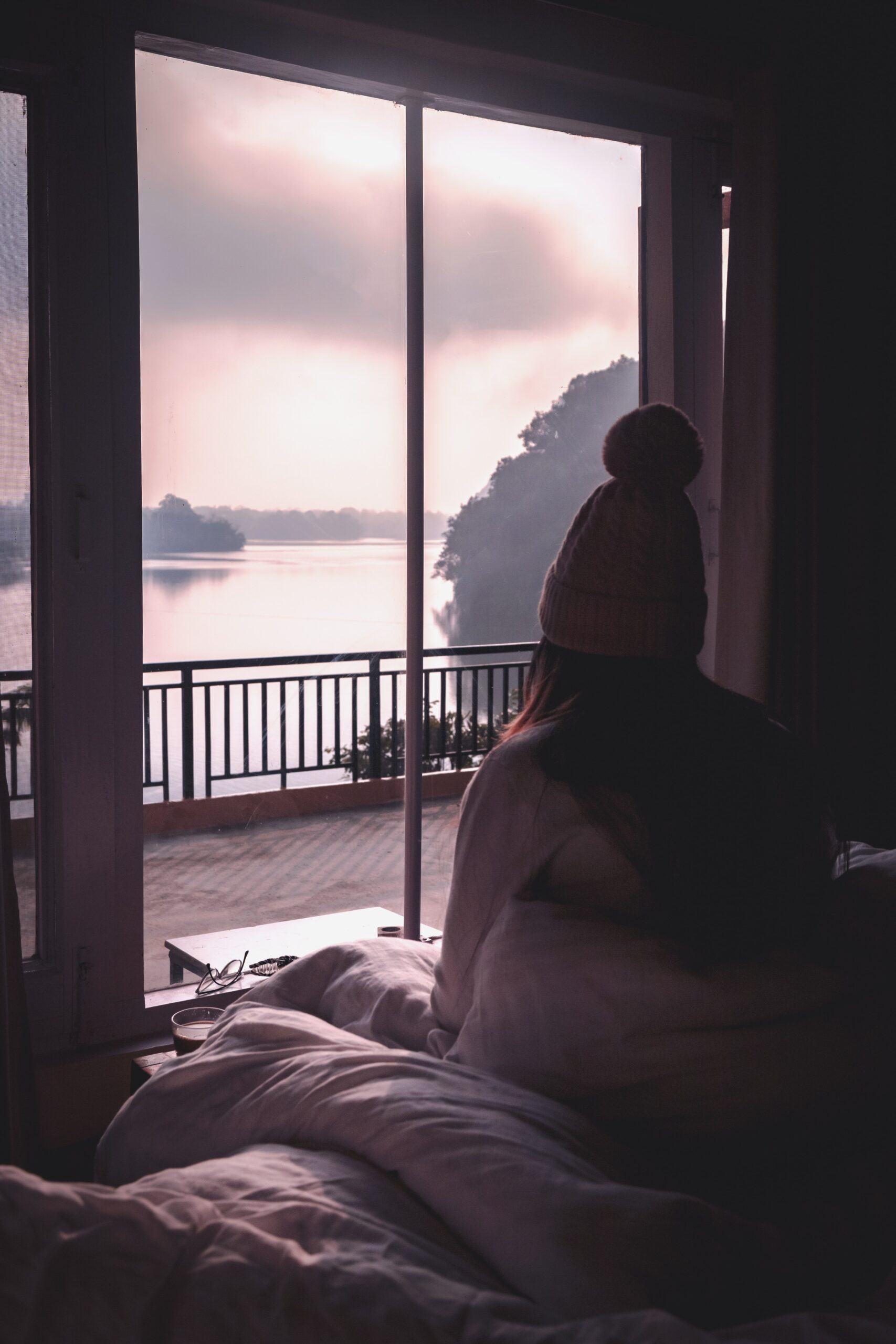 Woman sits with her back to the camera on a bed, looking outside her window past a balcony to a large misty river. 