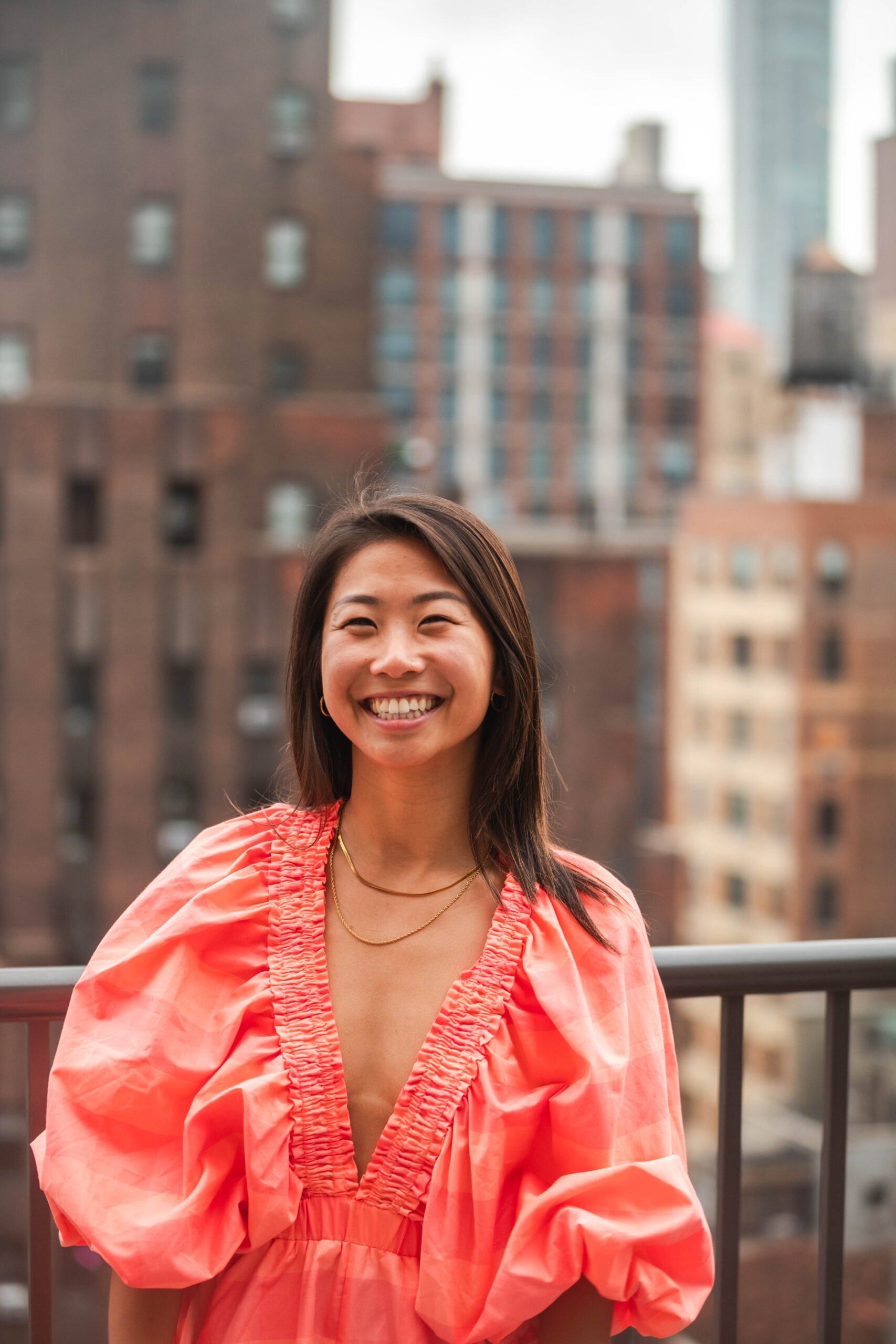 A portrait of Chris Wang, a Chinese Canadian woman, smiling brightly on a rooftop with a cityscape of brick buildings and high-rises in the background. She is wearing a coral pink dress with puffed sleeves and a deep V-neckline, along with gold jewelry. Her dark brown hair is styled straight, framing her face. The natural lighting highlights her warm expression, creating a confident and joyful atmosphere.