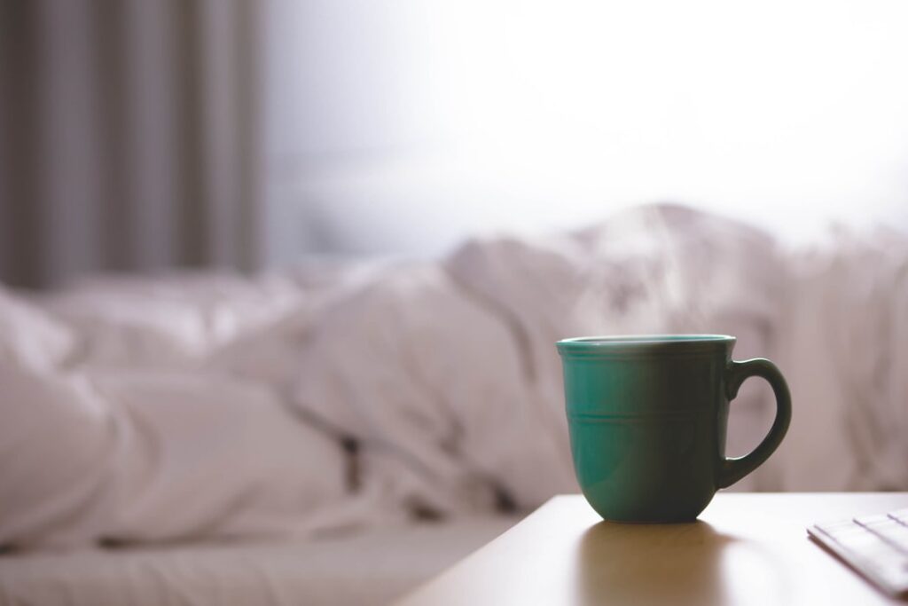 A green mug with a steaming beverage in the foreground. Crumpled up white bedsheets in the background.