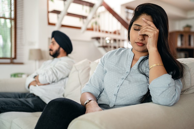 An Indian couple sitting on couch. Each person is showing body language that makes it clear that there's been a disagreement. One crosses their arms while the other grips their forehead.